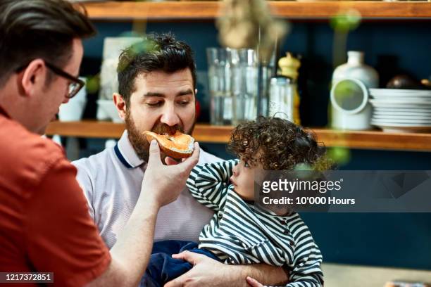 man with baby son being fed a bagel by husband - diverse family stock-fotos und bilder