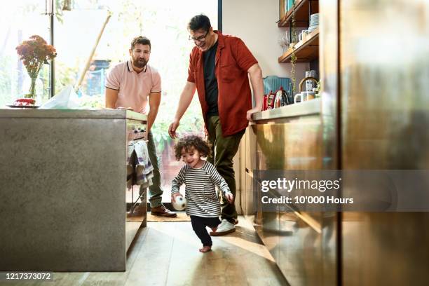 two fathers watching toddler playing with ball in kitchen - young family in kitchen stock pictures, royalty-free photos & images