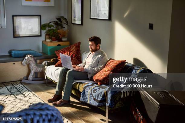 mature man with beard working from home on laptop - man sitting on sofa fotografías e imágenes de stock