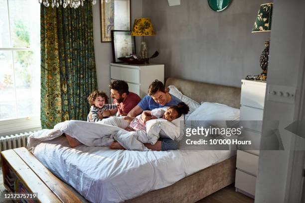 cheerful same sex couple tickling young children in bedroom - family on bed stock pictures, royalty-free photos & images