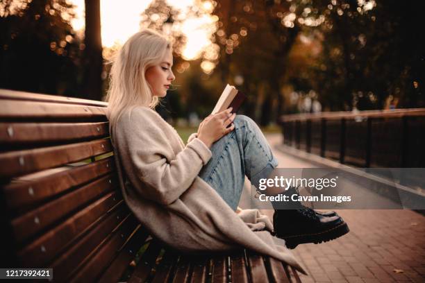 teenage girl reading book sitting on bench in park during autumn - girl wearing boots stock-fotos und bilder