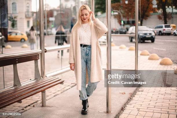 portrait of smiling young woman standing on bus stop in city - girl wearing boots stock-fotos und bilder