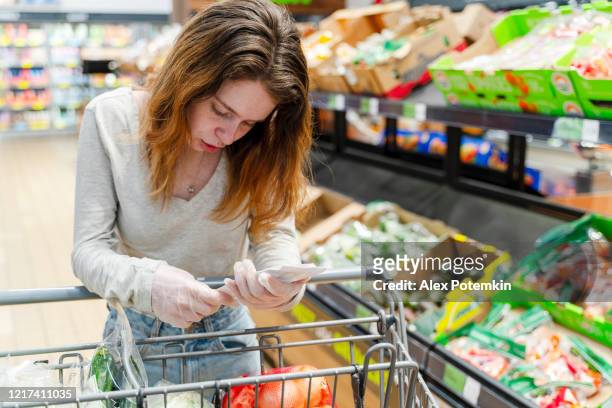 práctica de compras segura durante un brote de pandemia viral. una joven con guantes comprando verduras en una tienda, buscando en una lista de compras. - panic buying fotografías e imágenes de stock