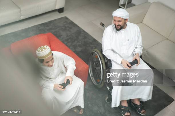 arabic man in wheelchair at home playing video games with son - covid persons with disabilities stock pictures, royalty-free photos & images