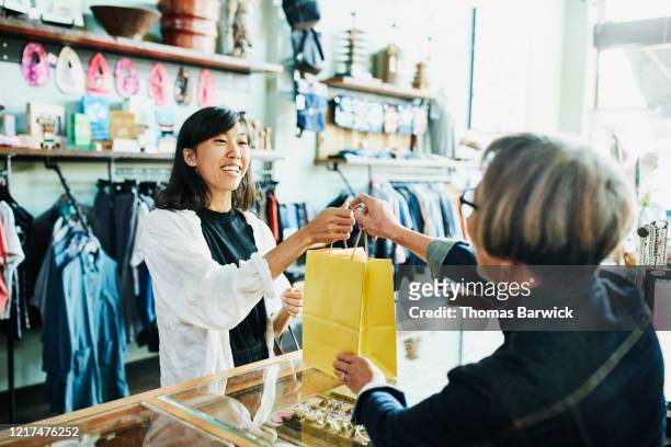 shop owner handing bag to smiling client after shopping in clothing boutique - retail place stock pictures, royalty-free photos & images