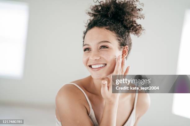 glimlachende jonge vrouwen die moisturiser op haar gezicht toepassen - glowing stockfoto's en -beelden