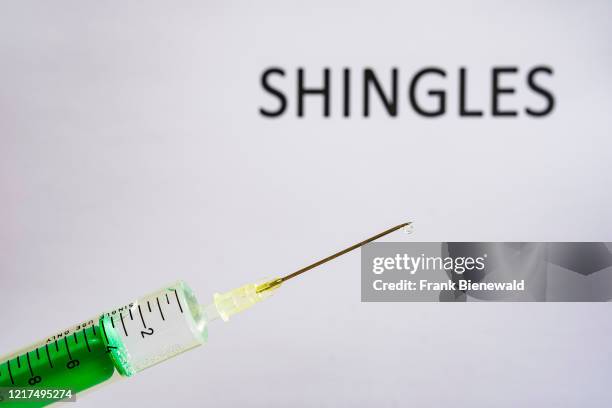 This photo illustration shows a disposable syringe with hypodermic needle, SHINGLES written on a white board behind.