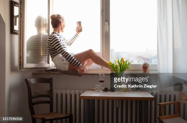woman sitting on a windowsill, video chatting with family member - quarantäne stock-fotos und bilder