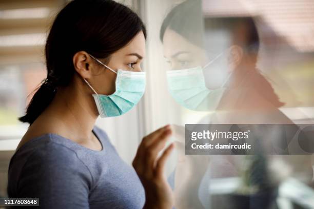 portrait of sad young woman with face protective mask looking through the window at home - quarantine stock pictures, royalty-free photos & images