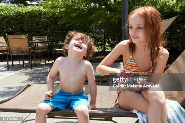 little boy and girl sitting outside in their swim suits on a lounge chair together. they are smiling and laughing and the little girl is taking something out of a little red bag. - schwimmbeckenrand stock-fotos und bilder