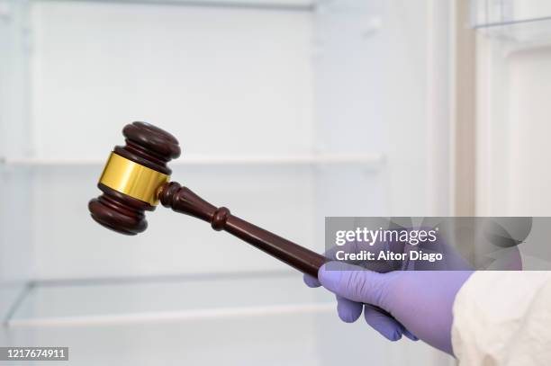 human hand wearing protection gloves with a judges gavel. empty fridge in the background. justice and coronavirus concept. - legal proceeding stock-fotos und bilder