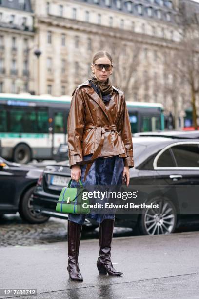 Guest wears sunglasses, a brown shiny jacket, a scarf, a blue skirt, a green bag, leather pointy boots, outside Ann Demeulemeester, during Paris...