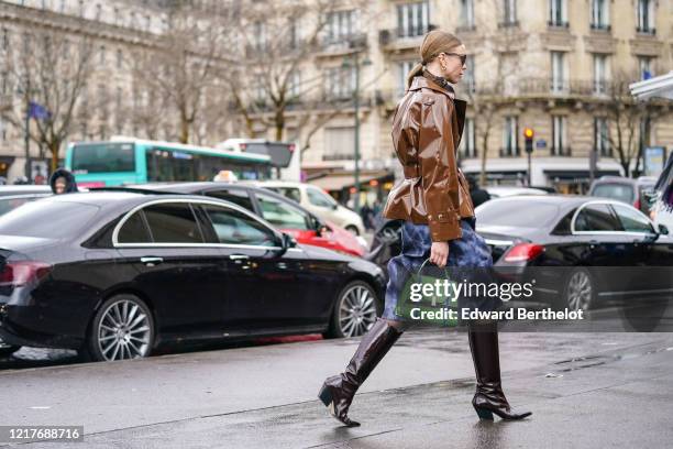 Guest wears sunglasses, a brown shiny jacket, a scarf, a blue skirt, a green bag, leather pointy boots, outside Ann Demeulemeester, during Paris...