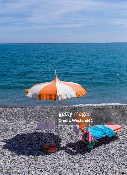 france, alpes-maritimes, cagnes-sur-mer, beach umbrella and deck chair on rocky coastal beach with clear line of horizon over mediterranean sea in background - provence alpes cote dazur stock pictures, royalty-free photos & images