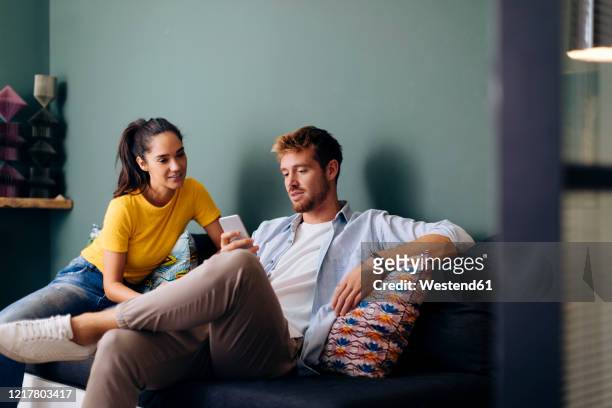 young couple sitting on the couch at home looking at smartphone - couple smartphone stockfoto's en -beelden