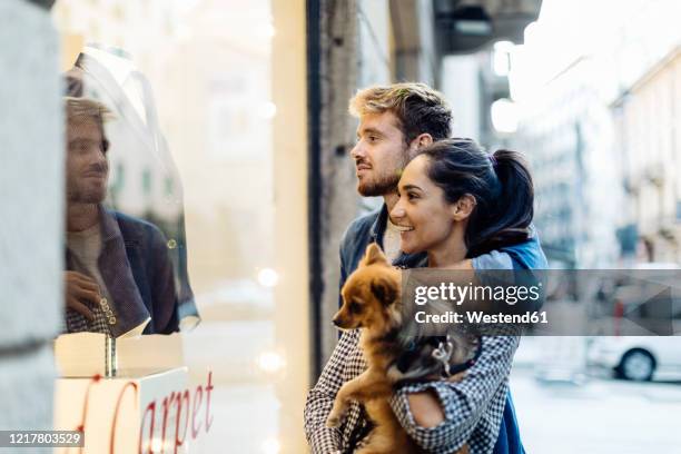 young couple with dog looking in shop window in the city - window shopping stock-fotos und bilder