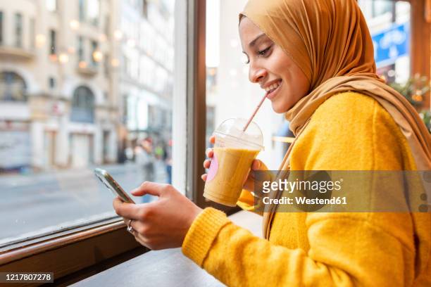portrait of happy young woman with smoothie and smartphone in a cafe - yellow smoothie stock pictures, royalty-free photos & images
