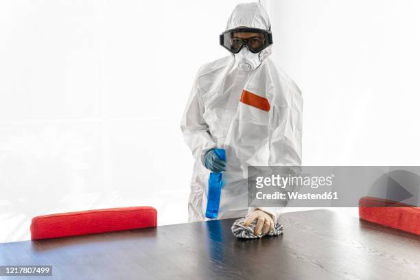 woman wearing protective clothes, sanitizing her home, wiping table - protective suit stock pictures, royalty-free photos & images