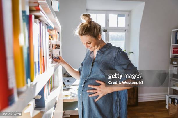 pregnant woman standing at bookshelf at home - preganant woman stockfoto's en -beelden