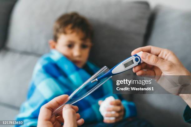 mother's hand taking digital thermometer while sich son waiting on couch in the background - febre imagens e fotografias de stock