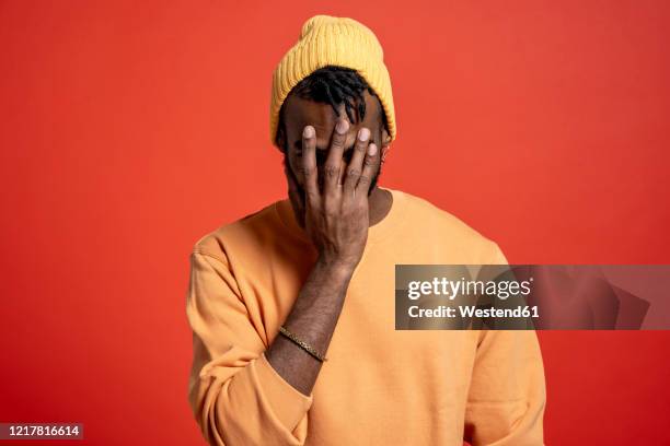 young man covering his face in front of orange wall - pessimisme stock pictures, royalty-free photos & images