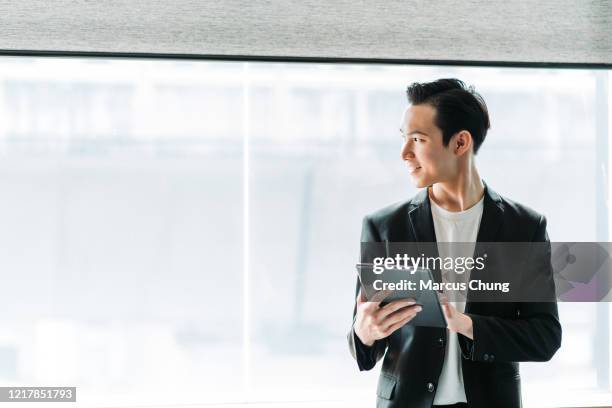 asiatische chinesische junge lächelnde geschäftsmann mit einem digitalen tablet und schauen weg in seinem büro - chinese businessman stock-fotos und bilder