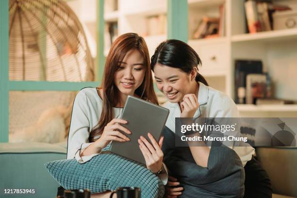 two asian chinese smiling businesswoman sitting together on sofa chatting, using digital tablet during tea break at indoor office relaxation area - facial expression girl office stock pictures, royalty-free photos & images