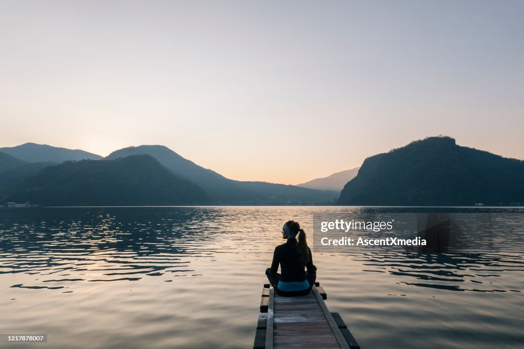 Woman sits on edge of dock over lake and watches sunrise over mountains and forest