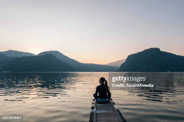 woman sits on edge of dock over lake and watches sunrise over mountains and forest - ponytail silhouette stock pictures, royalty-free photos & images