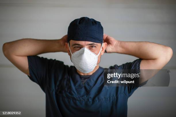 doctor in scrubs and face mask portrait during lockdown fastening mask looking at camera - doctor arms raised stock pictures, royalty-free photos & images