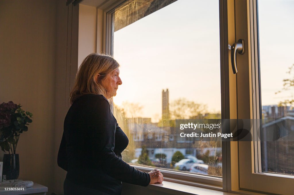 Senior woman looking out of her bedroom window at sunset