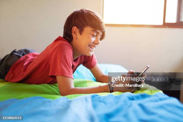 cute latinx hispanic boy smiling while texting on smartphone in his room - 12 year old cute boys stock pictures, royalty-free photos & images