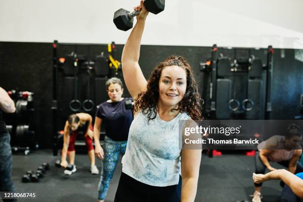 smiling woman doing one arm overhead dumbbell press during class in gym - woman gym stock pictures, royalty-free photos & images