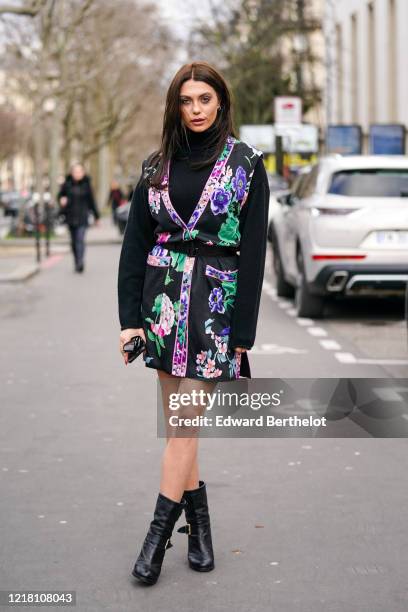 Heloise Agostinelli wears earrings, a black turtleneck pullover, a black green and purple floral print kimono dress, a belt, black leather boots,...
