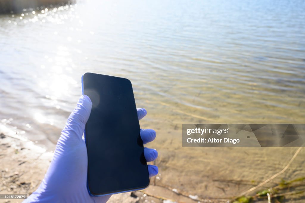 Human hand with protection globes holding a mobile phone a beach´s lake. Germany.
