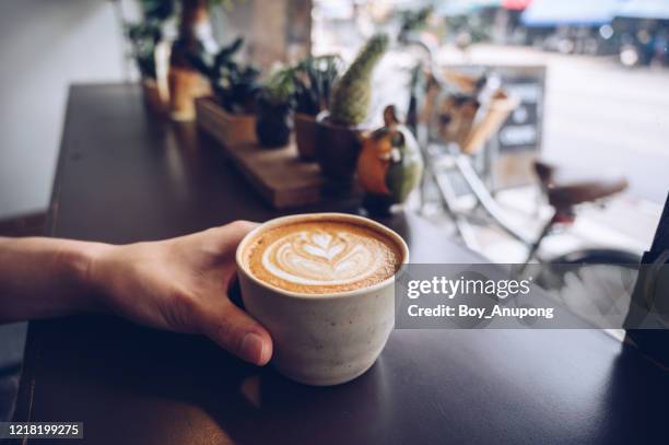 cropped shot of someone hand with a cup of hot latte coffee on the table nearly window. - cafe latte stockfoto's en -beelden