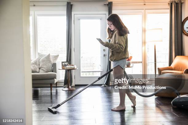 teenage girl vacuuming living room while using smartphone - beautiful barefoot girls - fotografias e filmes do acervo