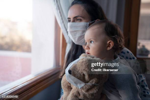bonita niña y madre whit máscara protectora de pie junto a la ventana con su oso de peluche - máscara quirúrgica fotografías e imágenes de stock