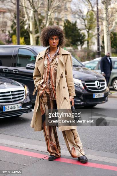Guest wears a light brown trench coat, a black bag, a brown and beige pyjama striped shirt, flare pants, leather shoes, outside Leonard Paris, during...