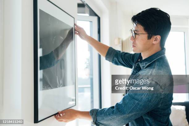 young man hanging picture on wall at home - hängen stock-fotos und bilder