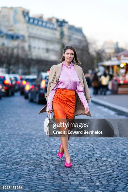 Landiana Cerciu wears a beige long trench coat, a pink silky shirt, an orange silky lustrous skirt, a white bag, neon pink pointy shoes, outside...