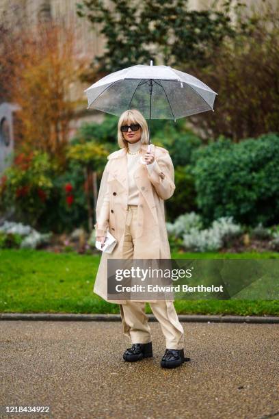 Xenia Adonts wears sunglasses, a white turtleneck pullover, a golden chain necklace, a beige clear long coat, beige pants, black shoes outside Maison...