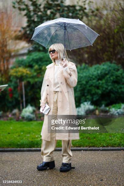 Xenia Adonts wears sunglasses, a white turtleneck pullover, a golden chain necklace, a beige clear long coat, beige pants, black shoes outside Maison...