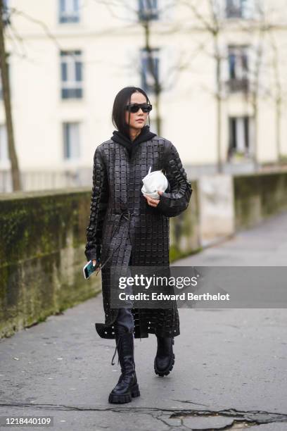 Yoyo Cao wears sunglasses, a black hoodie sweater, a black woven leather long coat, a black and white leather bag, leather shoes, outside Mugler,...