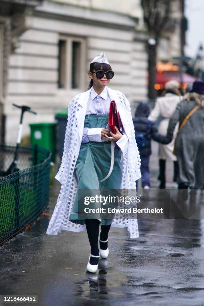 Leaf Greener wears a small hat, sunglasses, a white shirt, a white long coat with several holes, a green off-shoulder dress, black tights, white...