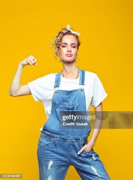 zuversichtliche junge frau in dungarees heben faust, porträt auf gelbem hintergrund - jeans latzhose frau stock-fotos und bilder
