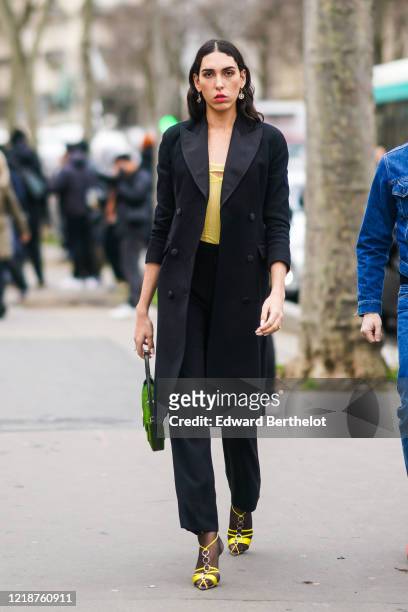 Guest wears earrings, a black long coat, a yellow top, black pants, yellow shoes, a green bag, outside Mugler, during Paris Fashion Week - Womenswear...