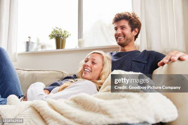 laughing young couple watching tv on their living room sofa - wrapped in a blanket stock pictures, royalty-free photos & images