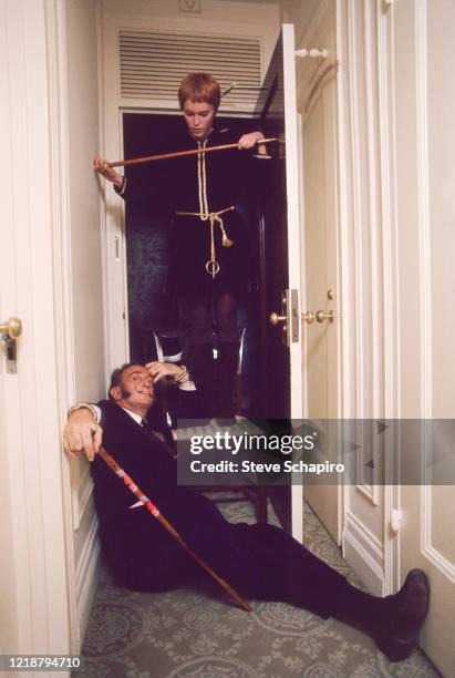 In an open doorway, American actress Mia Farrow stands on a chair looking down at Spanish Surrealist artist Salvador Dali , who sprawls on a hallway...