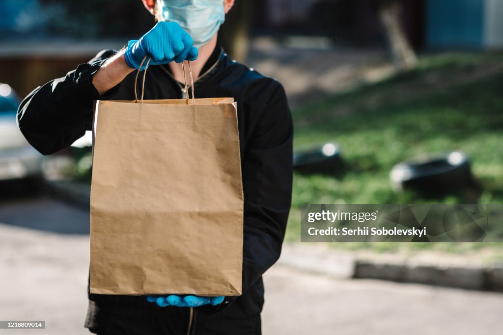 Courier in protective mask, medical gloves delivers takeaway food.  Employee hold cardboard package. Place for text. Delivery service under quarantine, 2019-ncov, pandemic coronavirus, covid-19.